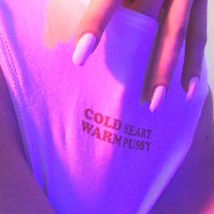 May include: Pale pink underwear with the text "COLD HEART WARM PUSSY" printed in brown. The image is lit with a purple hue.