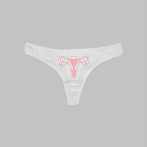 May include: White thong underwear featuring a pink illustration of a uterus. The underwear is made from a soft material and has a low-rise cut. The pink illustration is centrally placed on the front.