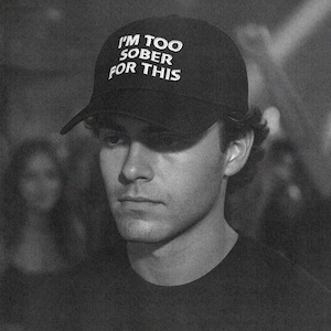 Pode incluir: Boné de beisebol preto com o texto "I'M TOO SOBER FOR THIS" em branco. O boné é usado por uma pessoa com uma camiseta preta. A imagem é em preto e branco.