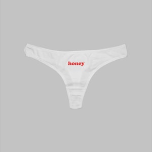 May include: White thong underwear featuring the word "honey" in red lettering. The underwear is made from a soft, lightweight material and has a classic thong design.