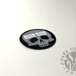 May include: A black and gray patch with a skull design and the text "SS" above the skull. The patch is oval-shaped and has a black border. The words "DARK SIDE DOJO" are printed in a circle.