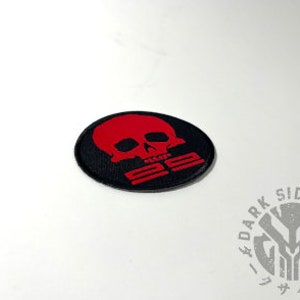 May include: A black, oval-shaped patch featuring a red skull design with the text "25". The patch is embroidered with a detailed design. The logo for "Dark Side Dojo" is visible in the lower right corner.