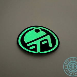 May include: A glowing green and black patch with a unique design. The patch features a circular shape with a stylized symbol in the center. The text "DARK SIDE DOJO" is visible on a smaller circular patch in the lower right corner.