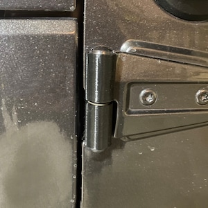 May include: Close-up of a black metal door hinge with a silver finish. The hinge is attached to a gray car door.