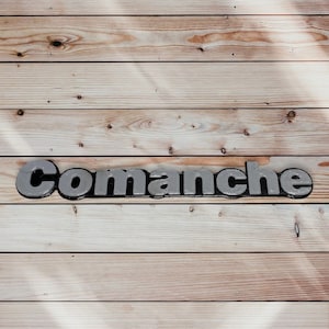 May include: The word "Comanche" is spelled out in silver 3D letters on a light brown wood background.