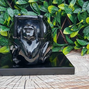 Op de afbeelding: Een zwart, glanzend, keramisch beeld van een aap die op een zwarte houten basis zit. De aap houdt zijn handen omhoog naar zijn hoofd en zijn ogen zijn gesloten.