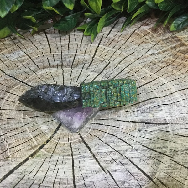 May include: A dark gray arrowhead with a green and black patterned stone wrapped around it. The arrowhead and stone are sitting on a purple amethyst crystal on a wooden surface.