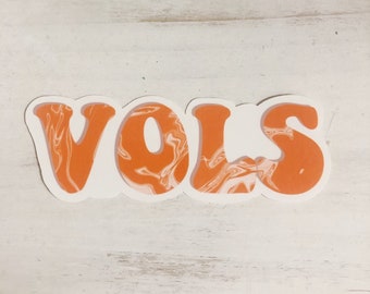 Könnte beinhalten: Ein weißer Aufkleber mit dem Wort "VOLS" in orangefarbenen Buchstaben im Retro-Stil. Die Buchstaben haben ein marmoriertes orange-weißes Muster. Der Aufkleber befindet sich auf einer weißen, verwitterten Holzoberfläche.