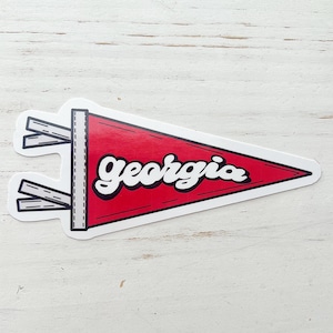 May include: A red pennant-shaped sticker with the word "Georgia" in white script. The sticker has a black outline and a white border. The pennant has a black and white design on the left side.