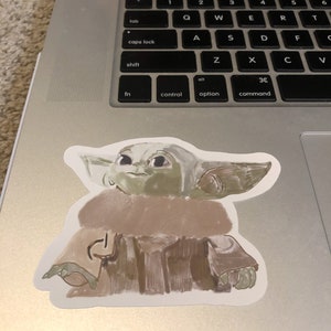 Op de afbeelding: Een sticker met een gestileerde illustratie van een personage met grote oren en een bruine mantel. De sticker is geplaatst op een zilveren laptop, het toetsenbord is zichtbaar. Het gezicht van het personage is groen.