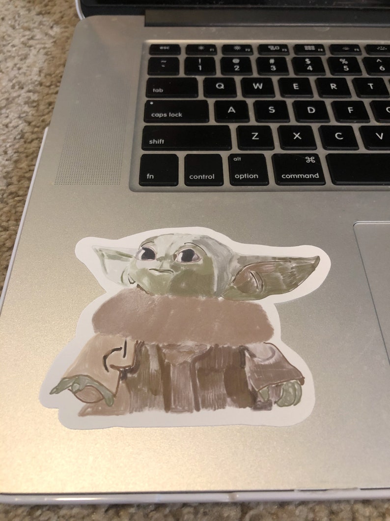 Op de afbeelding: Een sticker met een gestileerde illustratie van een personage met grote oren en een bruine mantel. De sticker is geplaatst op een zilveren laptop, het toetsenbord is zichtbaar. Het gezicht van het personage is groen.