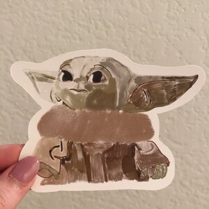 Op de afbeelding: Een sticker met een gestileerde illustratie van Grogu, ook bekend als Baby Yoda, uit The Mandalorian. Het kunstwerk gebruikt tinten bruin, groen en beige. Het personage heeft grote oren en een neutrale uitdrukking.