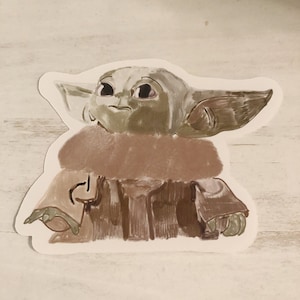 Op de afbeelding: Een sticker met een gestileerde illustratie van Grogu, ook bekend als Baby Yoda, uit The Mandalorian. Het kunstwerk toont Grogu in tinten groen, bruin en beige, met grote ogen en opvallende oren. De sticker heeft een witte rand.
