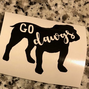 May include: A black silhouette of a bulldog with the words "GO dawgs" in white script. The image is on a white background, likely a sticker or decal, and is set against a speckled gray surface.
