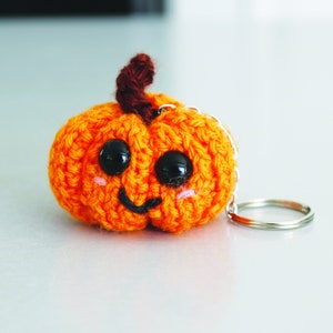 May include: A crocheted orange pumpkin keychain with black eyes and a smiling mouth. The keychain has a silver metal ring.