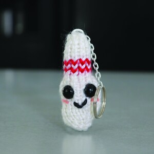 May include: White knitted bowling pin keychain with a red and white stripe around the middle. The pin has a smiling face with black eyes and a black mouth.