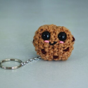May include: A brown crocheted cookie keychain with a smiling face, black eyes, and pink cheeks. The keychain has a silver metal ring and chain.