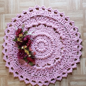 Könnte beinhalten: Ein runder rosa Häkelteppich mit einem kreisförmigen Design und einem dekorativen Rand. Der Teppich hat eine erhabene Textur und ist mit einem Blumenarrangement verziert.
