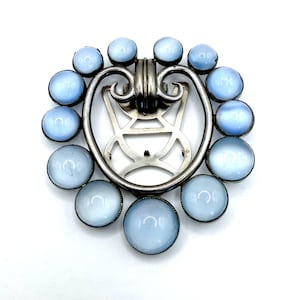 May include: A vintage silver-toned brooch with a central decorative element and a ring of light blue cabochon stones. The brooch has an ornate design with a clasp on the back. The stones are set in a silver-toned metal.