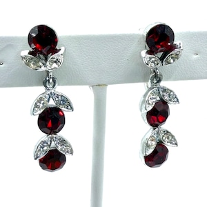 May include: A pair of silver-toned drop earrings featuring three faceted, deep red gemstones. Each earring has a floral design with clear, sparkling accents. The earrings are displayed on a white stand, showcasing their elegant design.