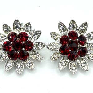 May include: A pair of flower-shaped earrings. Each earring features a cluster of dark red gemstones at the center, surrounded by clear, sparkling crystals and set in a silver-toned metal. The earrings are designed to resemble a flower.