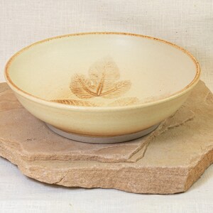 May include: A cream-colored ceramic bowl with a leaf design. The bowl is sitting on a light brown stone.
