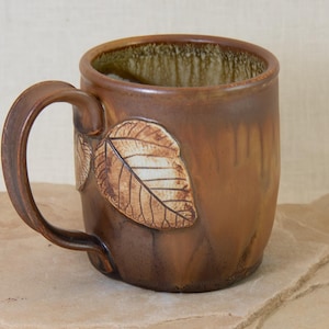 Puede incluir: Una taza de cerámica marrón con un diseño de hojas. La taza tiene un cuerpo ancho y redondeado y un asa.