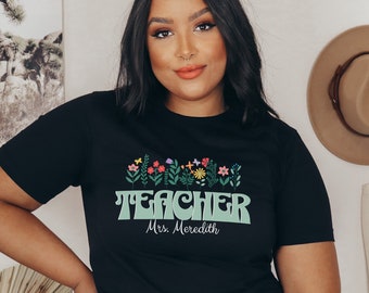 Pode incluir: Camiseta preta com um gráfico verde de "Teacher" com um design floral acima do texto. O texto "Mrs. Meredith" está abaixo do gráfico.
