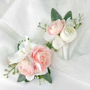 May include: Two pink and white flower boutonnieres with greenery. The boutonnieres are made of silk flowers and are perfect for a wedding or other special occasion.