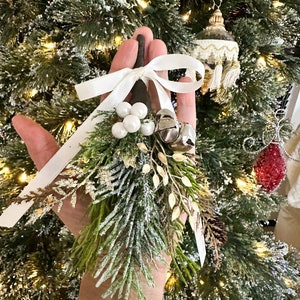 May include: A festive Christmas tree decoration with faux evergreen foliage, white berries, and silver bells. It is tied with a white ribbon. The ornament is held in front of a decorated Christmas tree.