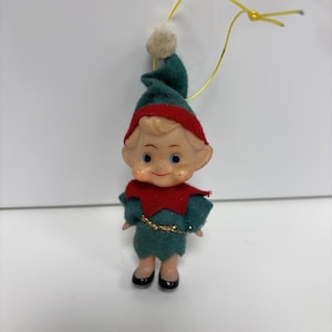 May include: A vintage Christmas elf ornament. The elf is dressed in a green felt outfit with a red collar and hat trim. The elf's face is painted with rosy cheeks and blue eyes. A gold-coloured string is attached for hanging.