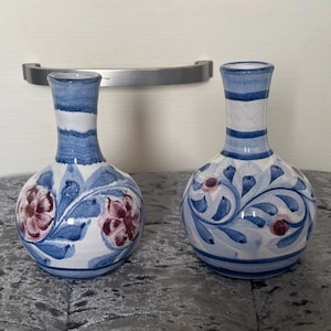 Può includere: Due vasi in ceramica con un design blu e bianco. I vasi hanno una base rotonda e un collo stretto. Un vaso presenta fiori rossi, mentre l'altro ha motivi floreali blu. I vasi sono su una superficie grigia.