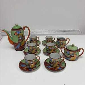 May include: A vintage tea set with a teapot, creamer, sugar bowl, cups, and saucers. The set features a green, orange, and brown color scheme with detailed artwork of a figure. The cups and saucers are small, and the set is likely for a small gathering.