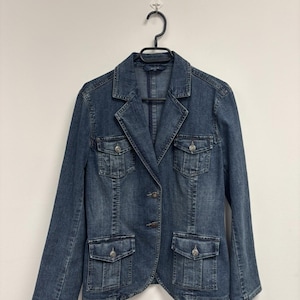 May include: A medium-wash denim blazer with a classic design. The jacket features a notched collar, four front pockets with button closures, and two buttons for fastening. The blazer is hanging on a black hanger against a white background.