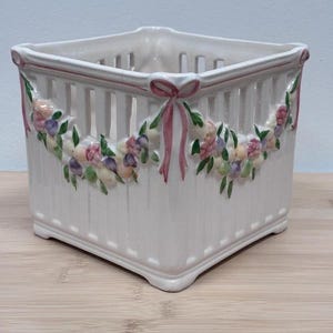 May include: A square white ceramic planter with a slatted design and a raised relief pattern of colorful flowers and green leaves draped in swags. The corners feature pink painted bows that connect the floral garlands around the exterior.