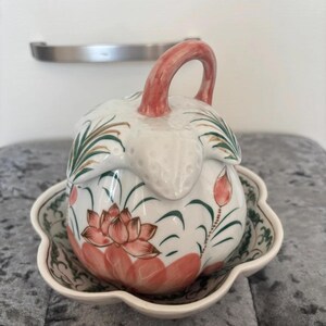 Puede incluir: Un recipiente de cerámica blanca con tapa y platillo a juego. El recipiente tiene forma de capullo floral, con un asa roja y diseños de flores de loto rosas. Acentos verdes y dorados decoran la tapa y el platillo.
