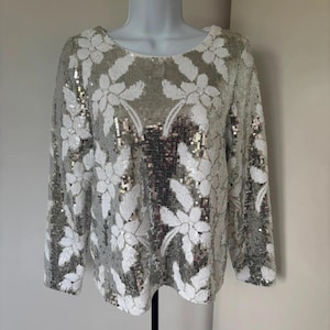 May include: Silver sequined long-sleeve top with white floral embroidery. The top is displayed on a mannequin. The sequins create a shimmering effect, and the floral design adds a touch of elegance. The top has a round neckline.