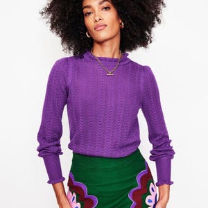Puede incluir: Una mujer con un jersey morado de manga larga con un patrón texturizado y un cuello alto con volantes. También lleva una falda verde con un diseño floral en tonos rosa, morado y marrón. La mujer lleva joyas doradas.