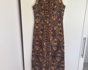May include: Sleeveless, floor-length dress with a high collar. The dress features a detailed pattern in shades of blue, brown, orange, and gold. The fabric appears to be a printed textile. The dress is hanging on a black hanger.
