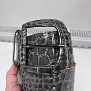 Puede incluir: Un cinturón ancho con un patrón brillante de relieve de cocodrilo en gris oscuro. El cinturón incluye una hebilla grande y redondeada a juego con un perno de metal oscuro pulido y una trabilla rectangular, con una textura en toda la correa.