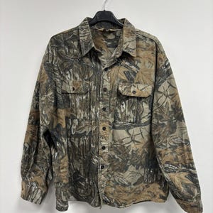 Può includere: Una camicia abbottonata a maniche lunghe con un motivo mimetico nei toni del marrone, verde e beige. La camicia ha due tasche sul petto e un colletto. Il tessuto sembra essere di peso medio.