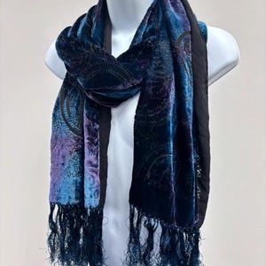 May include: A velvet scarf featuring a gradient of deep blue, purple, and black tones with a dark, intricate paisley pattern throughout. The scarf has a solid black border along the edges and is finished with long, dark blue fringe at both ends.