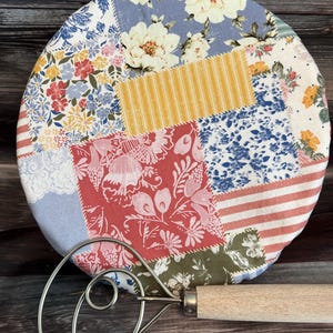 May include: A round fabric cover featuring a patchwork design of floral, striped, and lace patterns in shades of blue, red, yellow, and white, paired with a metal dough whisk that has a light-colored wooden handle positioned in the foreground.