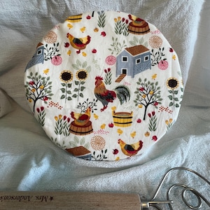May include: A white, circular kitchen item with a farm-themed pattern featuring roosters, chickens, sunflowers, and small houses. A wooden handled whisk is in the foreground. The text "Mrs. Anderson's" is visible on the wooden handle.