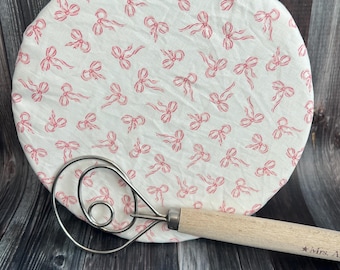 May include: A white, round bowl cover with a repeating pattern of pink bows. A stainless steel and wood whisk rests on the surface. The wooden handle is engraved with text.