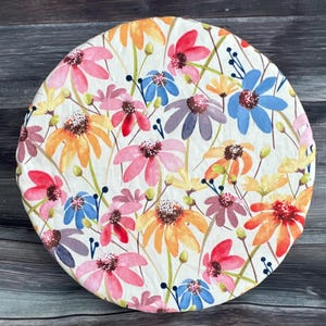 May include: A round, floral-patterned fabric item featuring a watercolor design. The design includes colorful flowers in shades of pink, orange, yellow, blue, and purple, set against a white background. Green stems and leaves are also present.