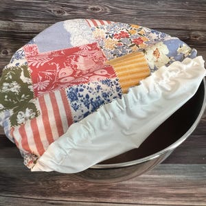 May include: A round fabric bowl cover featuring a patchwork design with various floral, striped, and solid patterns in shades of red, blue, yellow, green, and white. The cover has a white elasticized edge designed to fit securely over a metal bowl.