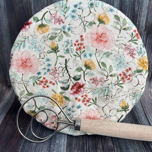 May include: A round, floral-patterned bread cover with pink, yellow, and blue flowers. A stainless steel dough whisk with a wooden handle rests in front of the cover. The cover is made of fabric.