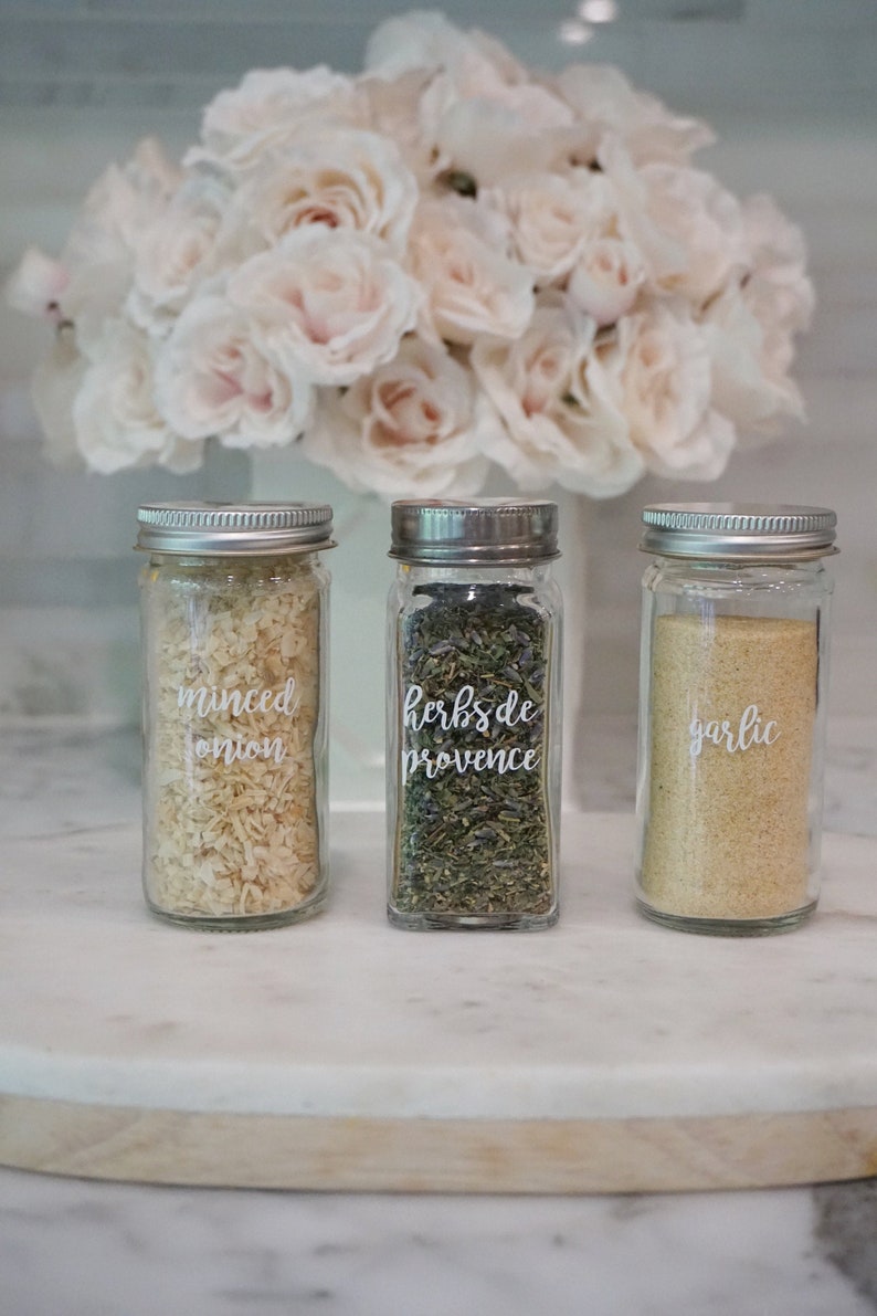 May include: Three glass spice jars with white labels on a white marble surface. The labels read "minced onion", "herbs de provence", and "garlic".