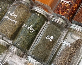 Könnte beinhalten: Nahaufnahme eines Gewürzregals mit verschiedenen Gewürzen in Glasbehältern. Die Behälter sind mit handgeschriebenen Etiketten versehen, darunter "Zimt", "Tajine", "Cayennepfeffer", "zerstoßener roter Pfeffer", "italienische Gewürzmischung", "Petersilie", "Dill", "Pfeffer", "Minze" und "Sesamsamen".
